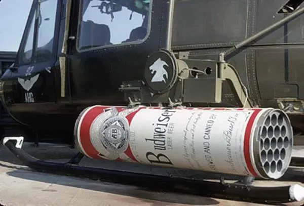 A military-style helicopter with a large, custom-made Budweiser beer can attached below its side, functioning as a humorous, multi-barrel launcher.