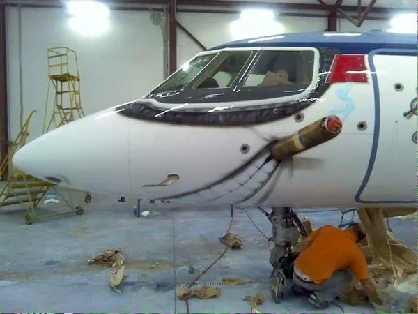 Close-up of an aircraft nose with an elaborate, humorous paint job depicting a grinning mouth biting a large cigar, being worked on by a mechanic.