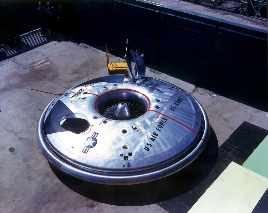 A top-down color photo of the Avrocar VZ-9, a silver disc-shaped experimental aircraft concept from the 1950s with "US AIR FORCE" and "US ARMY" markings, highlighting its unusual design as one of the crazy aircraft concepts that never truly flew.