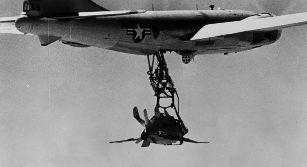 A black and white photo of the experimental McDonnell XF-85 Goblin "parasite fighter" dangling below its EB-29 mothership, showcasing a crazy aircraft concept that proved impractical for operational flight.