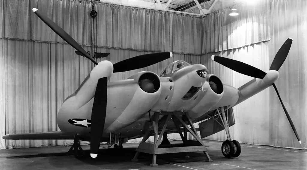 A black and white photo of the Vought XF5U "Flying Pancake" experimental aircraft mockup on display in a hangar, showcasing its unusual circular wing design and four large propellers, representing a crazy aircraft concept that never achieved full operational flight