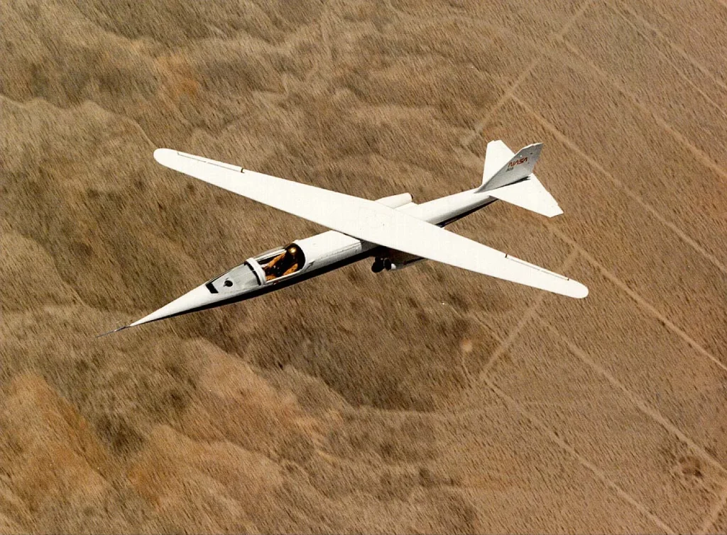 The white NASA AD-1 experimental oblique wing aircraft in flight over a brown, arid landscape, prominently displaying its single, slender wing swiveled at a sharp angle to the fuselage.