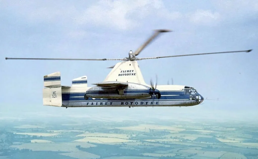 The Fairey Rotodyne experimental compound helicopter (XE521), featuring a white and blue livery with "FAIREY ROTODYNE" written on its side, in stable horizontal flight over a green, rural landscape, clearly showing its large main rotor and twin wing-mounted propellers.