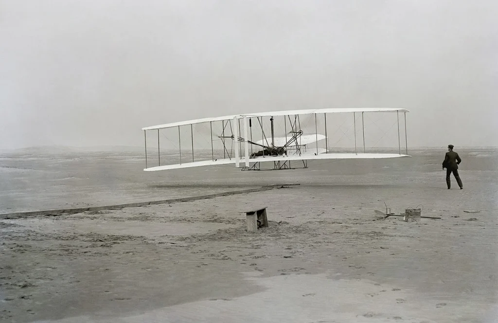 A black and white photo of the historic Wright Flyer during its first flight in 1903, with Orville Wright piloting the biplane and Wilbur Wright running alongside, marking the dawn of powered aviation.