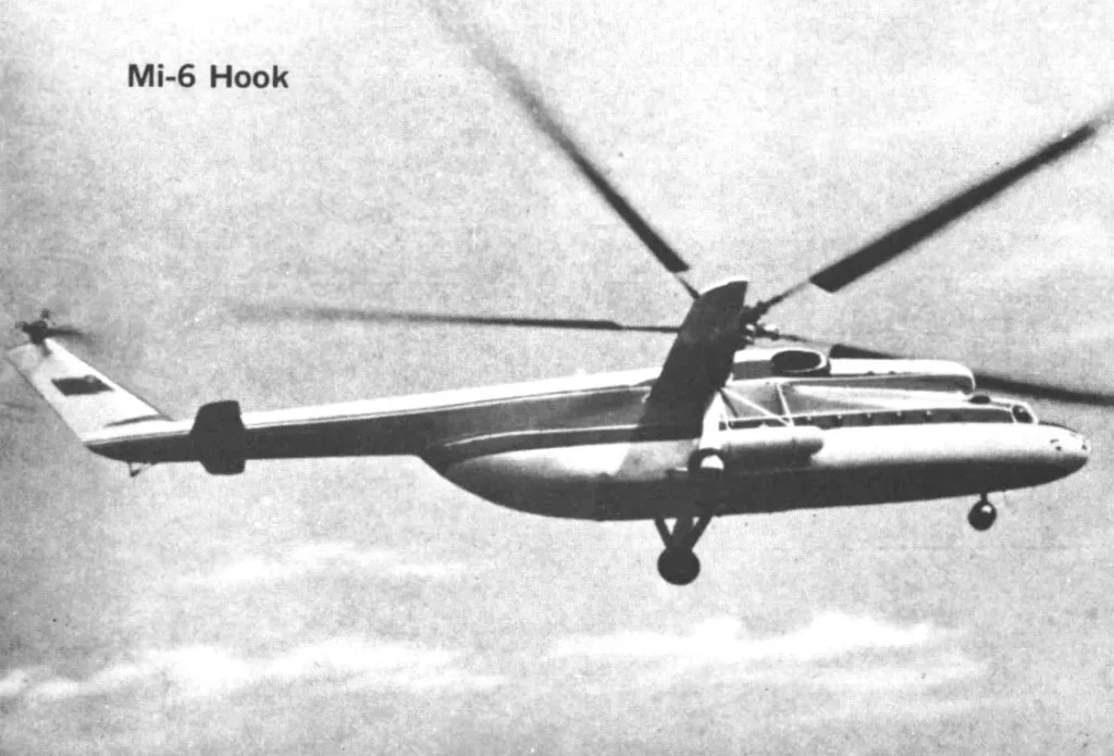 A black and white photograph of the Mil Mi-6 'Hook' heavy-lift helicopter in flight, seen from a side profile, highlighting its massive size, long fuselage, and single main rotor.