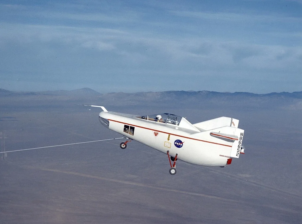 The white NASA M2-F1 experimental lifting body aircraft, nicknamed 'The Flying Bathtub', is seen in flight with its unique, wingless, half-cone shaped fuselage, red stripe, and NASA logo visible against a hazy sky with distant mountains.