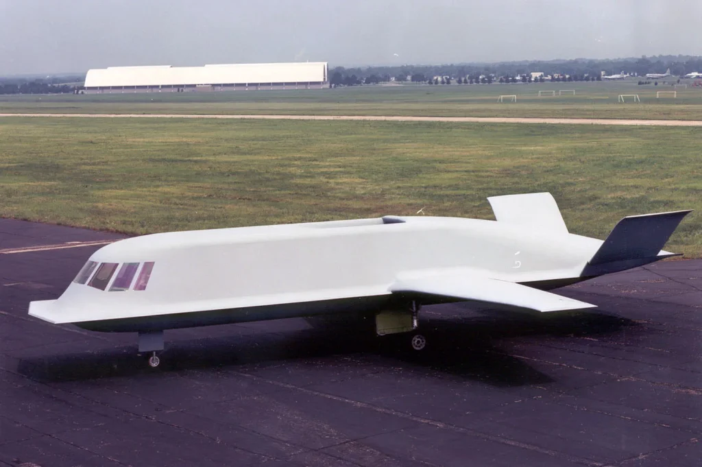 The Northrop Tacit Blue experimental stealth demonstrator, nicknamed 'The Whale', is shown parked on a tarmac with a large building in the background. Its distinctive, flat, gray, and highly angular fuselage lacks conventional wings and tail, giving it a futuristic and unconventional appearance.