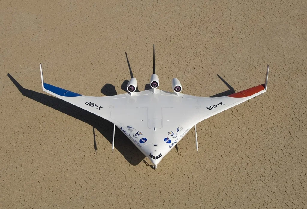 An aerial view looking down on the Boeing X-48B experimental blended wing body aircraft, with its sleek white, blue, and red manta-ray-like shape and three visible engines mounted on its upper surface, parked on a flat, tan surface.